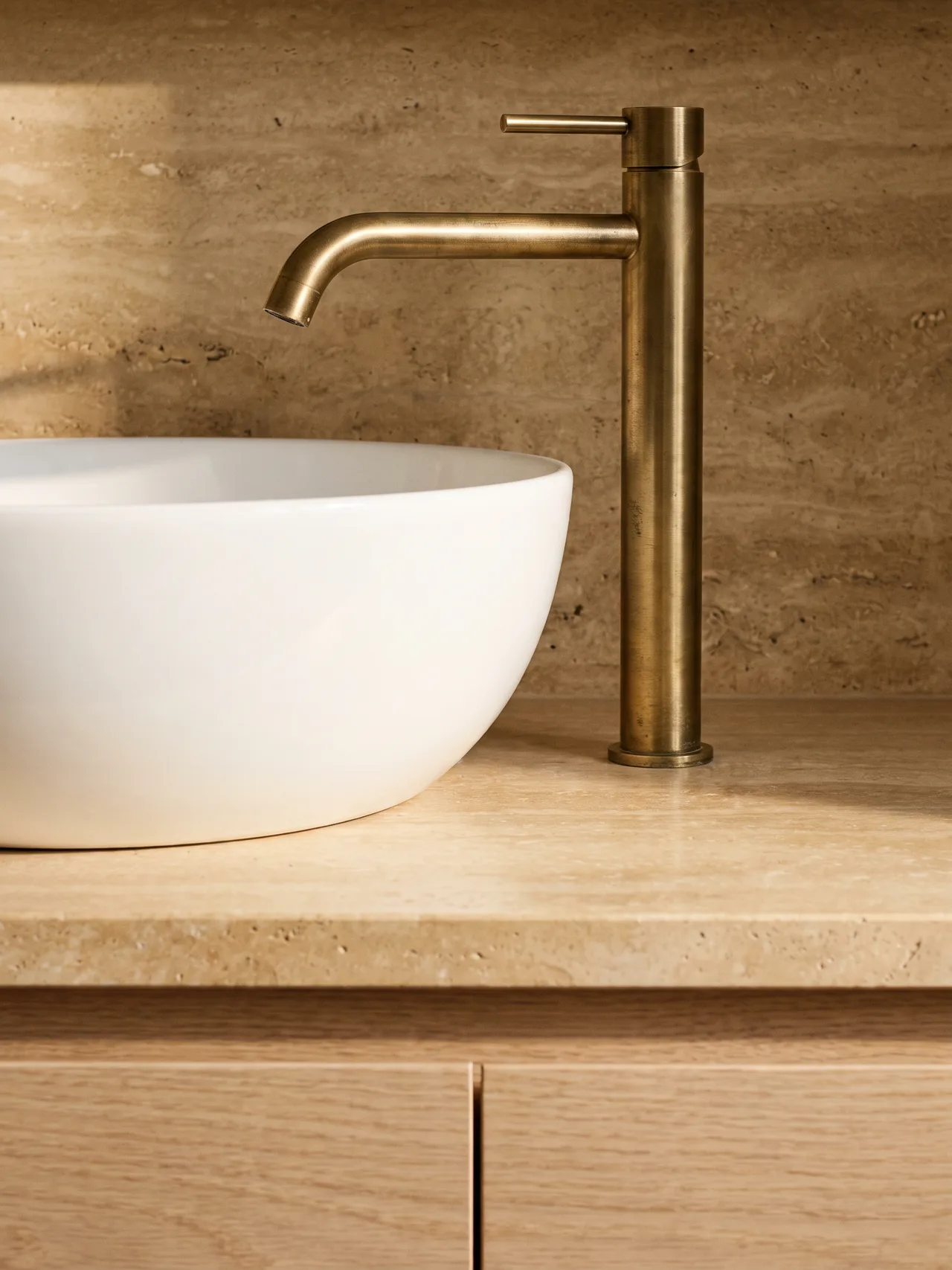 A white porcelain vessel basin and a tall aged brass mixer tap on a honey travertine countertop, with a travertine wall and pale oak drawers below, lit by morning light